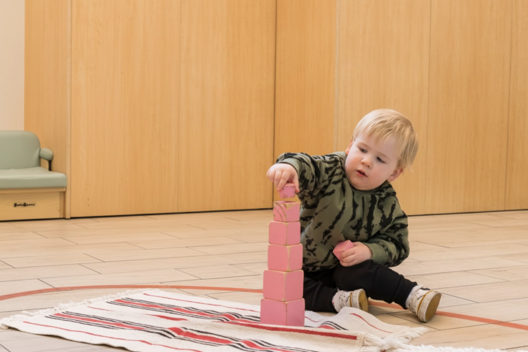 child on the floor stacking wooden pink cubes in order of size from biggest on the bottom to smallest at the top
