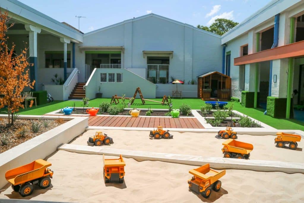 Well-equipped outdoor area at Belmore Montessori Academy with a sandbox filled with toy trucks, a lush green turf, and a wooden playhouse, framed by the centre's modern building facade.
