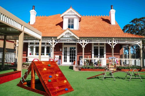 Charming outdoor area of Burwood Road Montessori Academy, with a wooden climbing ramp on green turf and a picturesque heritage-style building in the background under a clear blue sky.