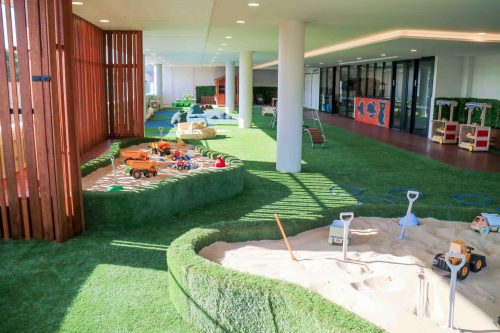 Peaceful indoor-outdoor play space at Crows Nest Montessori Academy with a sandbox full of toys, green turf, and an open area with educational activities, bathed in natural light