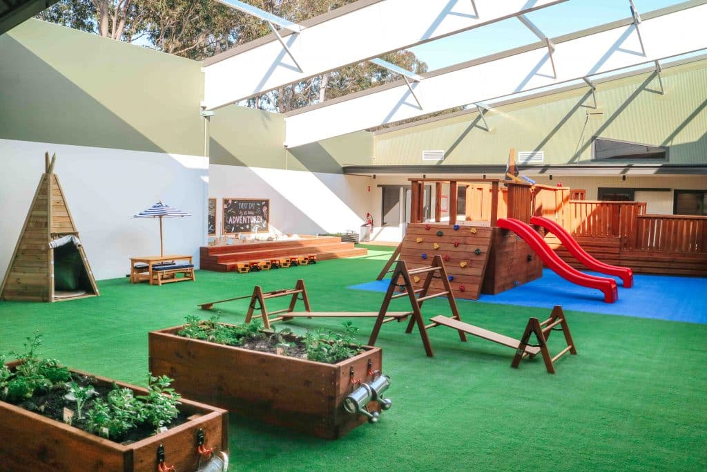 Spacious playground at Green Hills Montessori Academy with a tepee, raised garden beds, wooden play structures, and a bright red slide, all under an open-roof structure that bathes the area in natural sunlight
