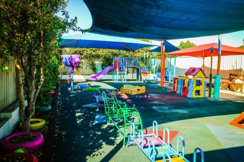 Shaded outdoor playground at Greenacre Montessori Academy with vibrant play equipment, colourful painted tyres, and a variety of activity areas on a resilient surface, offering a stimulating environment for children