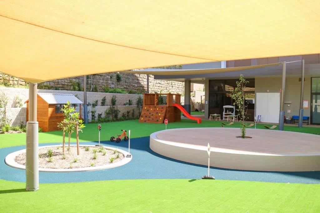 Contemporary outdoor play area at Lane Cove West Montessori Academy, with a large yellow sunshade, circular raised platform, wooden play structures, and lush plantings, offering a serene and engaging environment for children