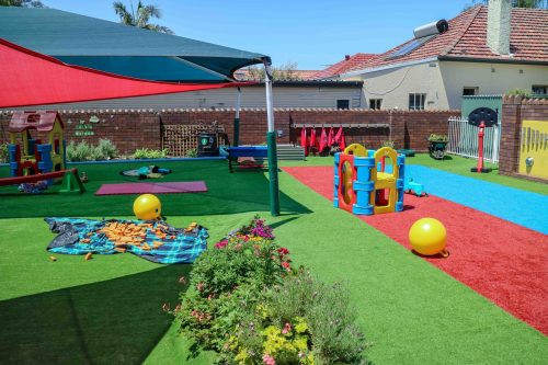 Dynamic outdoor playground at Croydon Montessori Academy with colourful safety surfacing, a variety of play equipment, shaded areas for comfort, and vibrant yellow balls, all designed for interactive play and learning