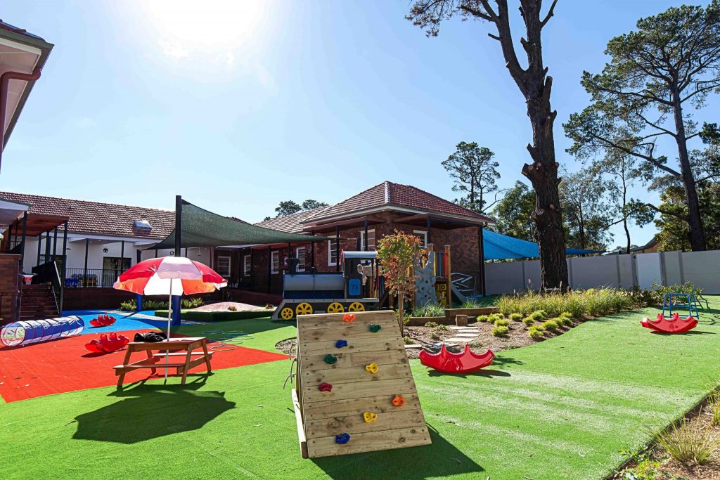 Sunny outdoor play area at Engadine Montessori Academy with a red and white umbrella, a climbing wall, and colourful playground equipment on artificial grass, complemented by a serene garden setting