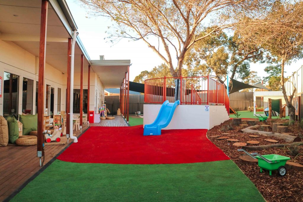 Outdoor area at Narellan Richardson Road Montessori Academy with a bright blue slide, lush green grass, red safety surfacing, and natural wooden elements, creating a vibrant and safe play space for children
