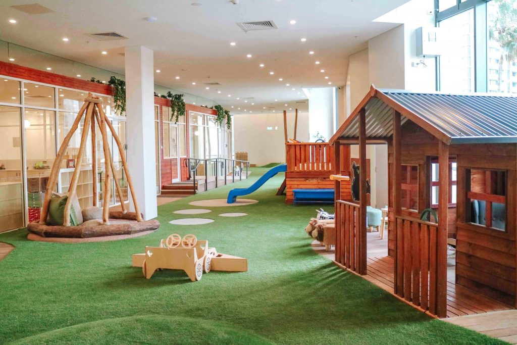 Inviting indoor play area at Newcastle Montessori Academy with a green grass floor, a tepee-like structure, a wooden playhouse, and a blue slide, all under a bright and spacious environment with natural lighting