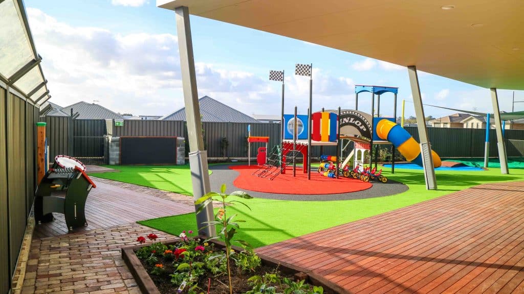 Spacious outdoor playground at Oran Park Montessori Academy, featuring a colourful play structure with slides, a racetrack, and a sheltered wooden deck, set within a well-landscaped area with a vibrant green lawn