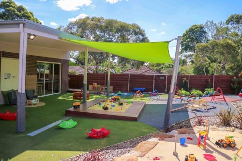 An outdoor play area at Turramurra Montessori Academy, with grass and play equipment like climbers and a sandpit, shaded by green sails, and enclosed by a wooden fence for safe play