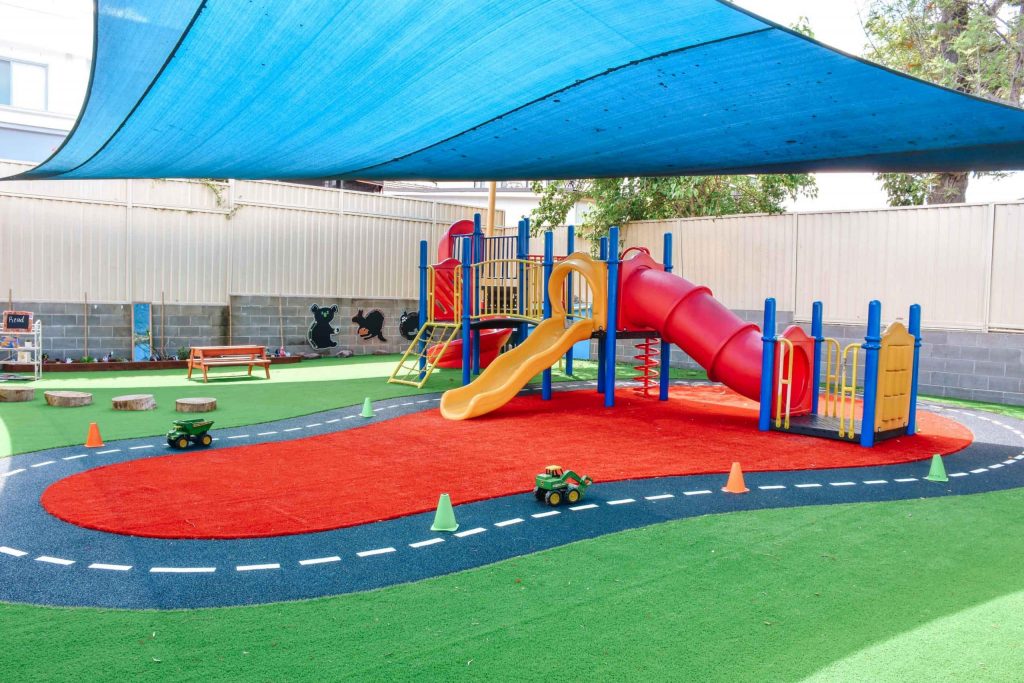 Colourful play area at Auburn Montessori Academy with a blue sunshade, red slide, miniature roadway, and play benches on turf