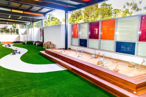 Modern outdoor play area at Carlingford Montessori Academy, with a sandpit and play scoops, green turf with a white winding path, and vibrant red and orange window panels