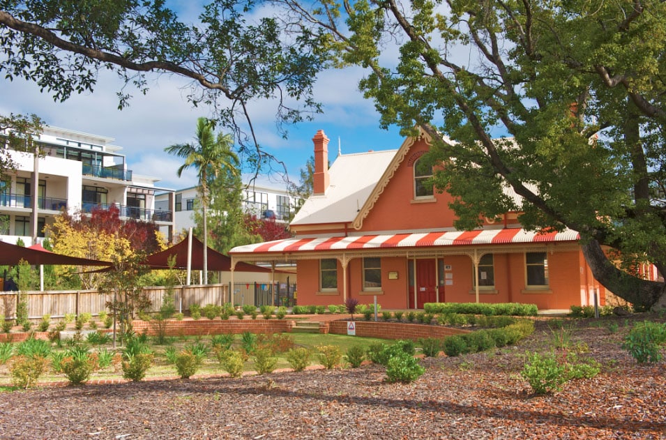 Historic red brick building at Castle Hill Montessori Academy, nestled among the greenery with modern buildings in the background, featuring striped awnings and a fenced playground shaded by mature trees