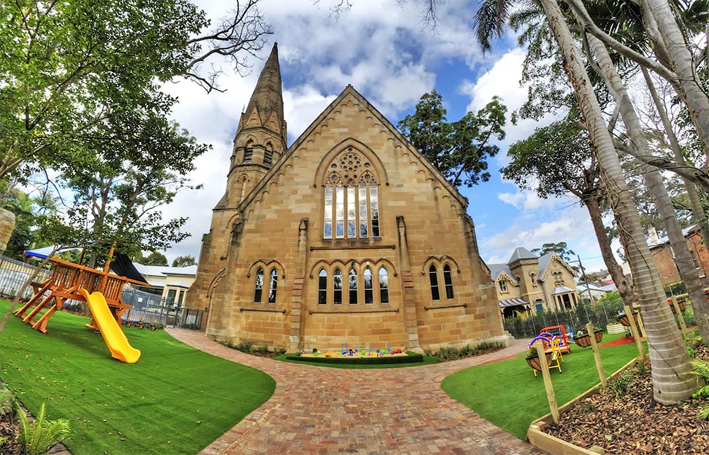Playground adjacent to a historic sandstone church at Glebe Montessori Academy, with a large yellow slide, play structures, and well-maintained green grass, creating a unique blend of heritage architecture and modern educational facilities