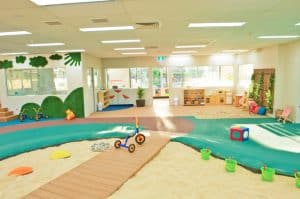 Large indoor playroom at Macquarie Park Montessori Academy with a creative play area featuring a sandpit, blue river-like carpet, and educational toys, accompanied by wall murals and natural light filling the space