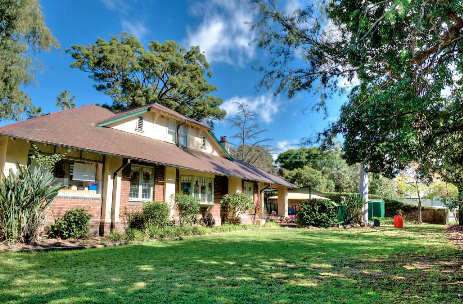 Historic, homely building of Strathfield Montessori Academy on Homebush Road, set amongst tall pines and lush greenery, presenting a warm and inviting environment for early learning with expansive lawns and a clear blue sky overhead