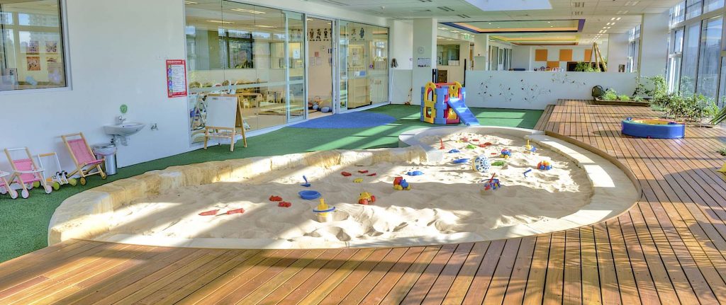 A bright indoor play space at Victoria Park Zetland Montessori with a circular sandpit, toys, and wooden decking. Glass-walled classrooms show a tidy, Montessori-style interior. Grass ensures safety, with ample natural light