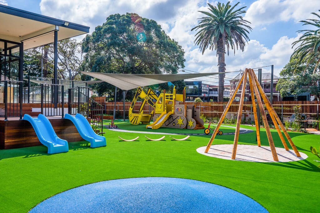 Cheerful playground at Burwood Park Montessori Academy featuring bright blue slides, a yellow excavator play structure, and a wooden swing set on green artificial turf under modern shade structures.