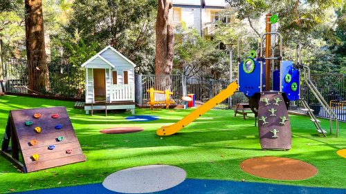 Shaded playground at Lane Cove Montessori Academy surrounded by natural greenery, featuring a climbing wall, playhouse, and a multifunctional play structure with slides, set on a vibrant green turf with colourful play spots