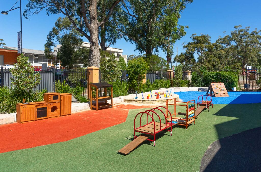 Vivid outdoor playground at Gymea Montessori Academy featuring imaginative play equipment like a wooden kitchen and climbing structures on safety surfacing, with mature trees providing natural shade