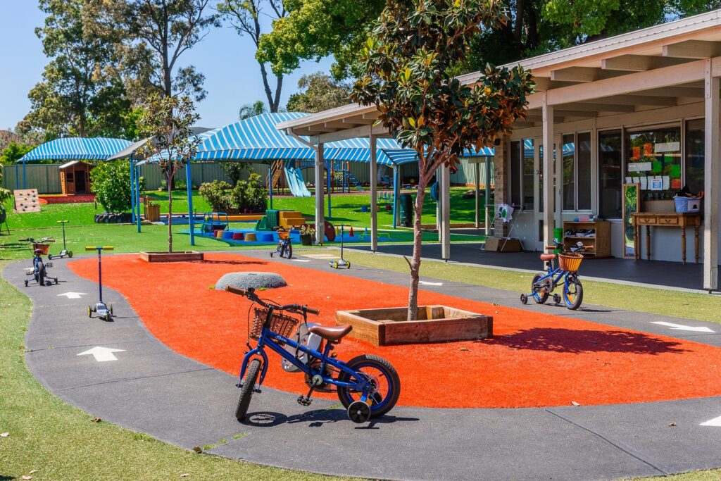 Lively outdoor playground at Smithfield Montessori Academy with a miniature road for bikes, red safety surfacing, green grass, and striped blue-and-white sunshades, encouraging active play in a secure, colourful environment