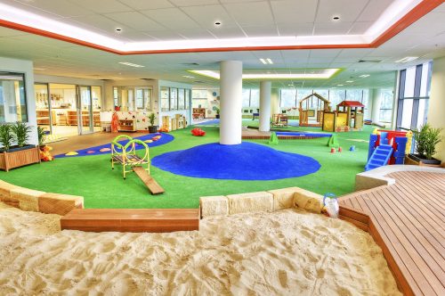 Spacious indoor play area at Bankstown Montessori Academy with a large sandpit, grass, central blue soft fall rubber, and various play stations including a wooden playhouse