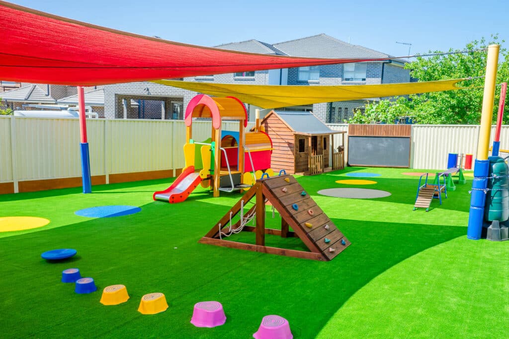 Colourful outdoor play area at Condell Park Montessori Academy, with multicoloured sunshades, a bright playground structure with a slide, wooden climbing ramp, and stepping stones on lush green grass