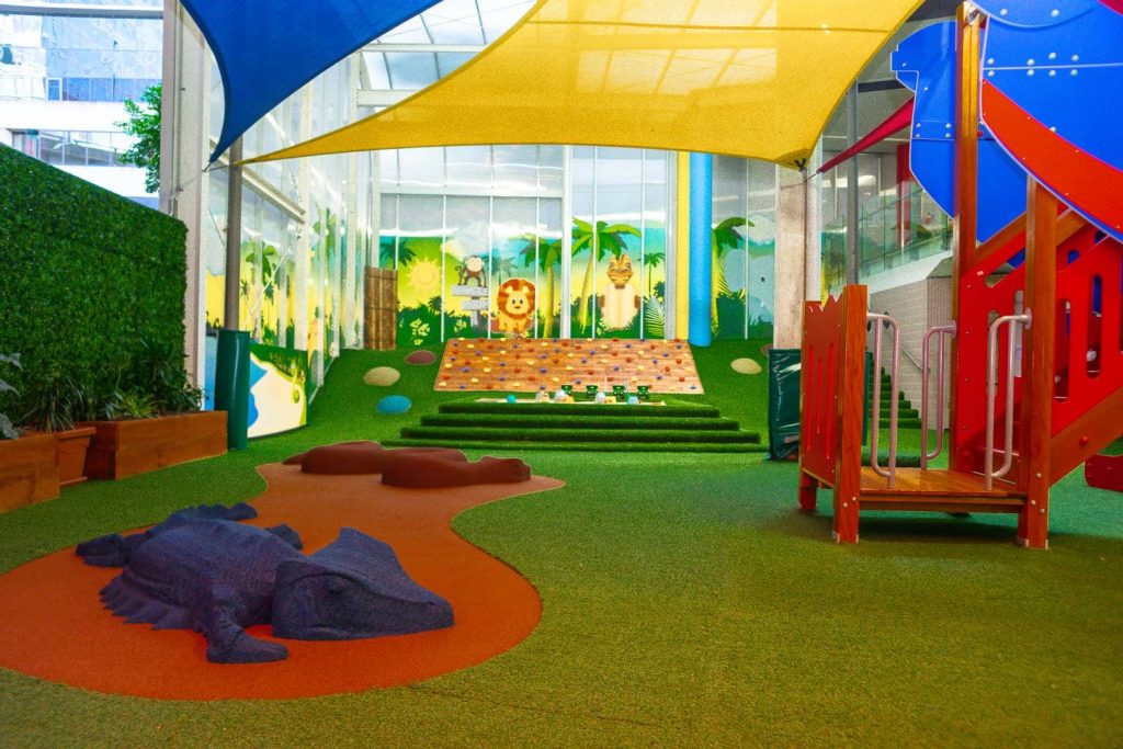 A vibrant and playful indoor play area at Barangaroo Montessori Academy, awash with colours under a canopy of yellow and blue shades. Features include a faux lizard and snake, a climbing wall, a jungle-themed mural, and a red and blue play structure, all designed to stimulate young minds and encourage active play
