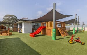 Spacious outdoor play area at Montessori Academy in Forest Hill, featuring a bright red slide, a wooden fort, a climbing wall with colourful handholds, and a trike track under a sun-protective shade sail
