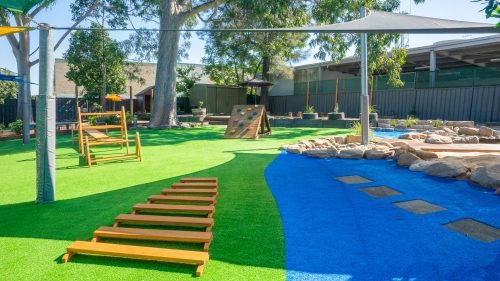 Lush outdoor play environment at Penrith Montessori Academy with a green lawn, wooden play structures, stepping stones across a blue stream-like feature, and natural boulders, shaded by mature trees and modern canopies