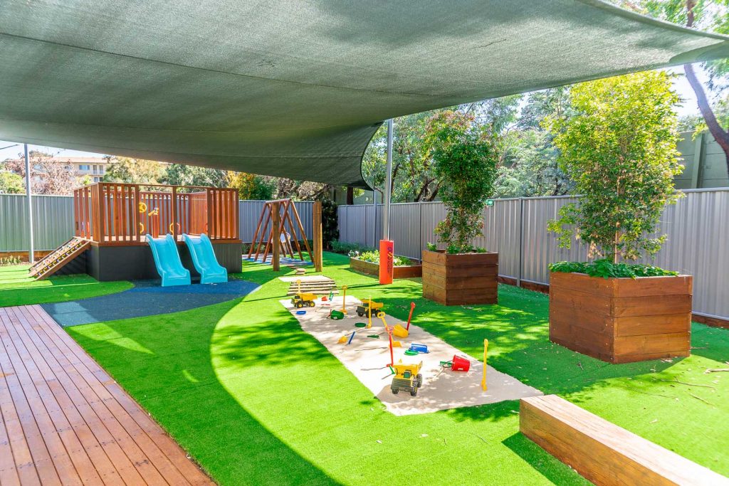 Sheltered outdoor play area at Queanbeyan Montessori Academy featuring bright blue slides, a climbing frame, large planter boxes with lush greenery, and various play toys scattered across the vibrant green artificial turf