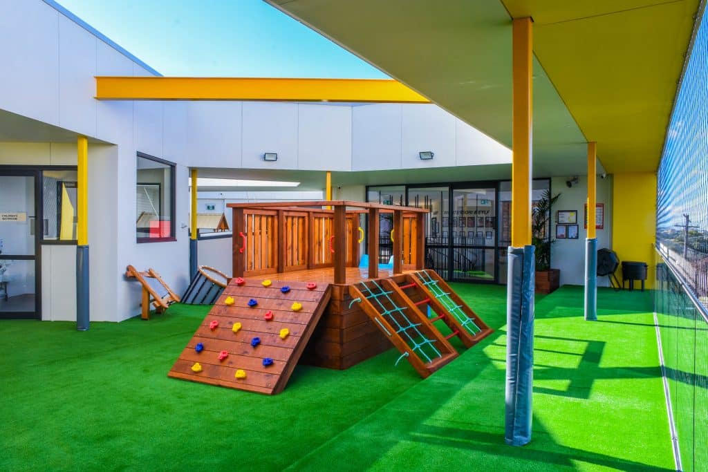 Sunny outdoor playground at Belfield Montessori Academy, featuring a wooden climbing structure with a rock wall on vibrant green turf, under a yellow and white awning, next to the academy's sleek, modern exterior