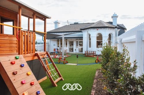 Interactive outdoor play area at Montessori Academy in Elsternwick, showcasing a wooden climbing structure with a slide, vibrant climbing holds, and a rope ladder, set against the backdrop of the centre's classic architecture and manicured lawns