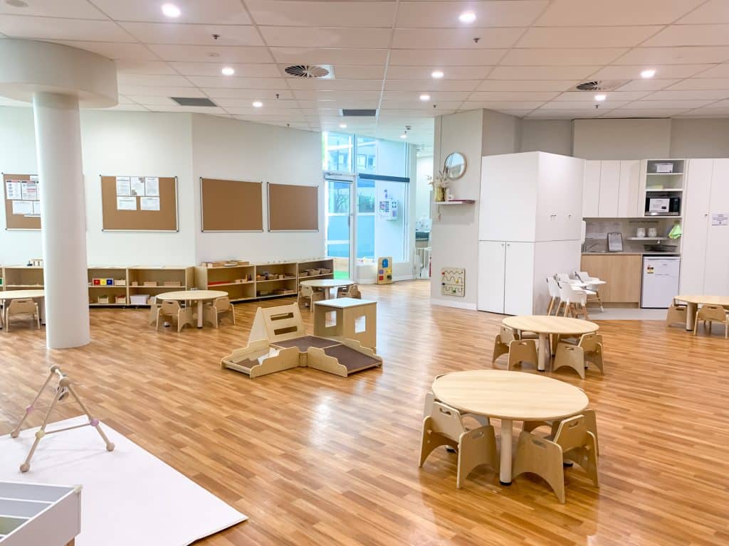 Clean and well-organised interior of Chatswood Montessori Academy with wooden furniture, learning materials on shelves, round tables for children, and a kitchenette area, illuminated by natural light from large windows