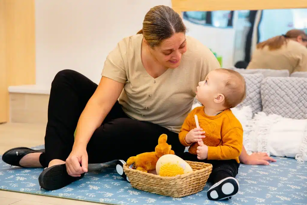 childcare educator and infant sitting on a mat, looking at each other and smiling