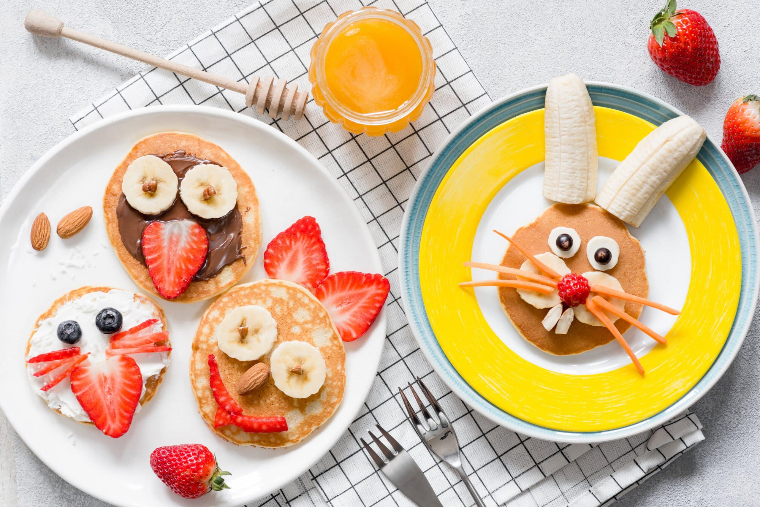 Healthy Easter-themed pancakes decorated to look like a bunny and chick, surrounded by fresh strawberries, bananas, and a dish of honey.