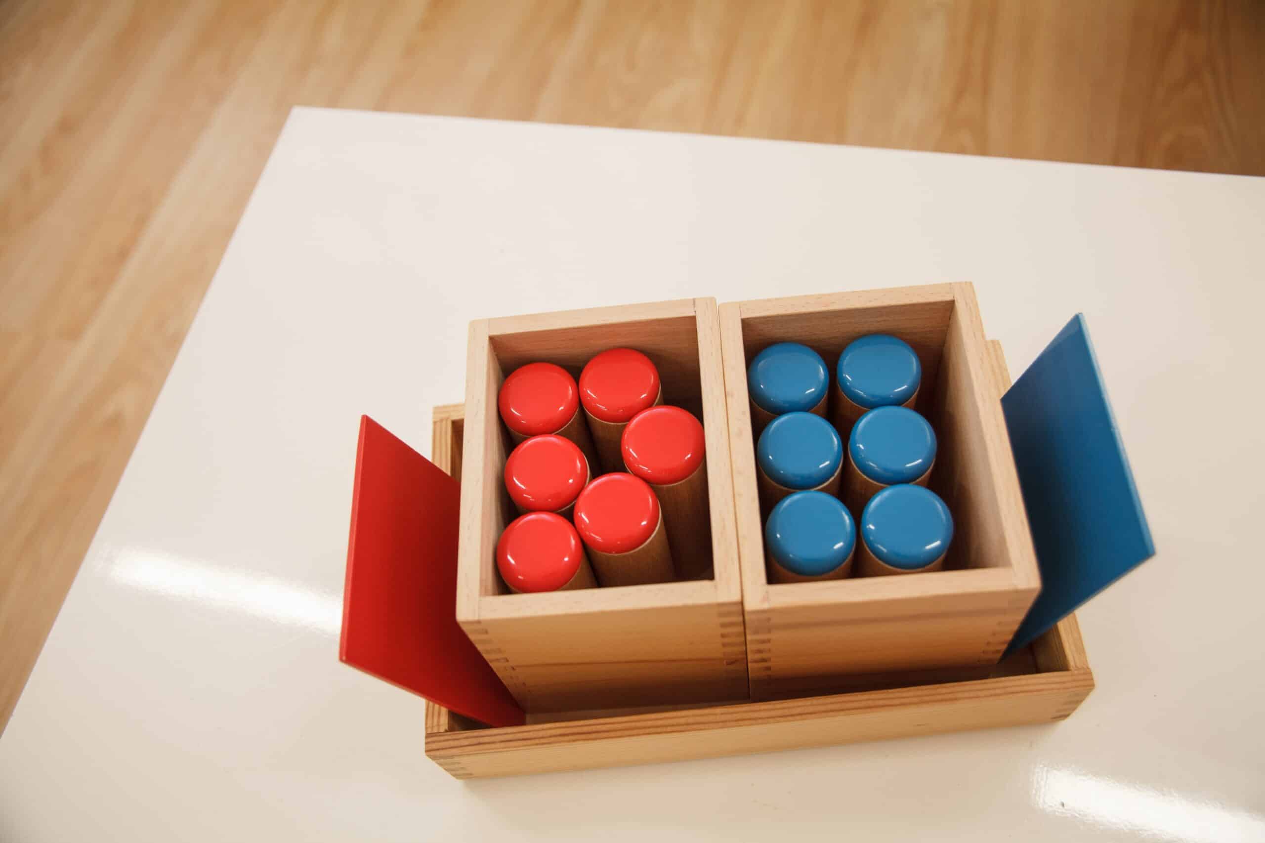 Wooden Montessori sound boxes with red and blue coloured lids on a white table.