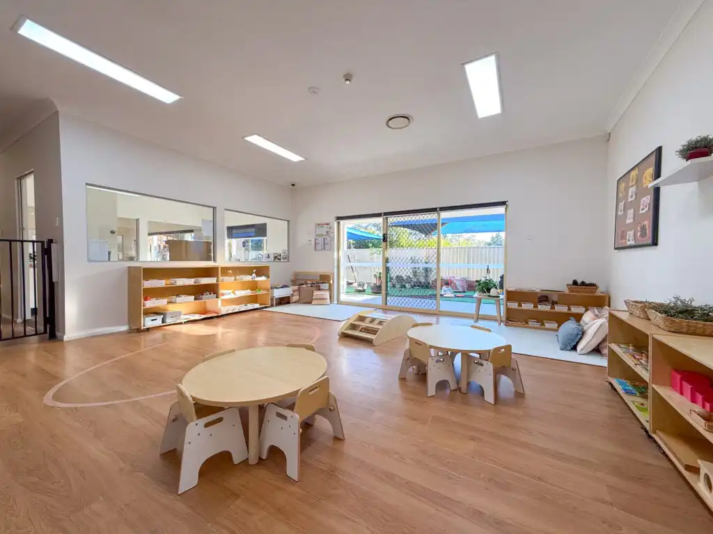 Spacious Montessori classroom at Auburn Albert Road childcare with round tables, learning shelves and large windows to outdoor area