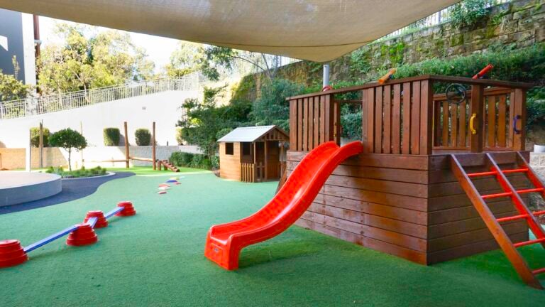 Outdoor playground at Montessori Academy’s Lane Cove West childcare, with a slide, playhouse and turfed play areas.