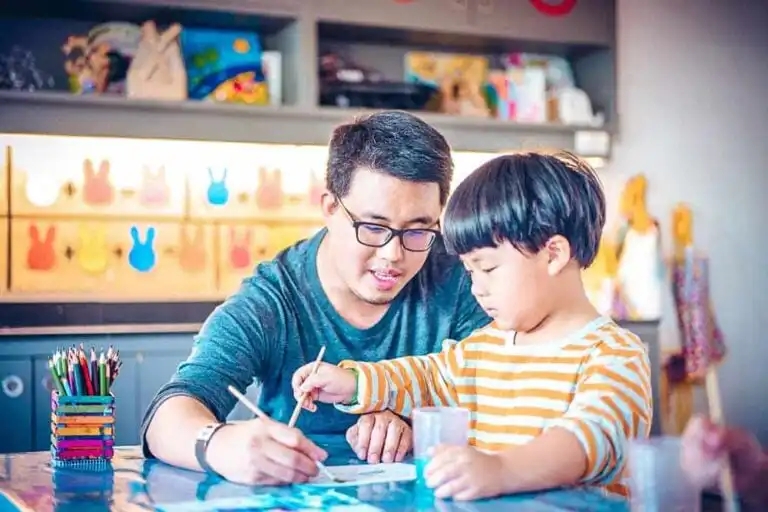 A father with glasses teaching his young son a painting activity.