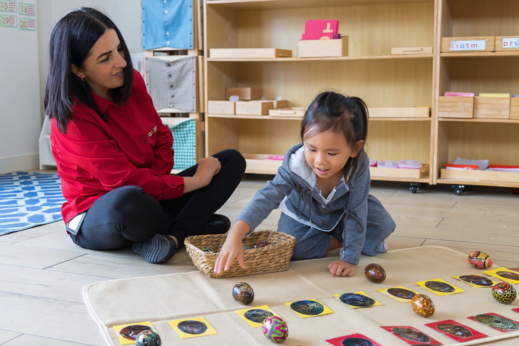In a classroom, an educator sits cross-legged on the floor, observing a young girl who is matching a small object to its corresponding image on a work mat.