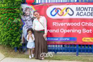 Pregnant mother and young daughter stand in front of red grand opening banner outside Riverwood Montessori Academy