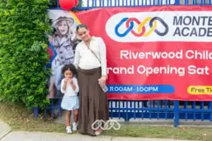 Pregnant mother and young daughter stand in front of red grand opening banner outside Riverwood Montessori Academy