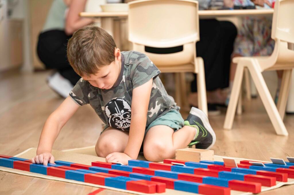A young child using Montessori red and blue number rods to explore early numeracy concepts and counting skills on a classroom floor.