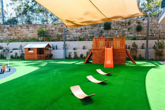 Shaded outdoor playground at Lane Cove West Montessori Academy featuring a timber climbing fort, slide, cubby house, balancing equipment, and open green play spaces.