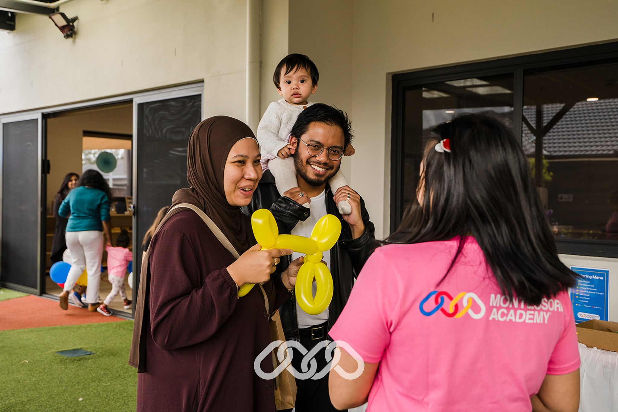 father holds daughter on his shoulders and stands next to wife in brown scarf talking to enrolment advisor in pink shirt