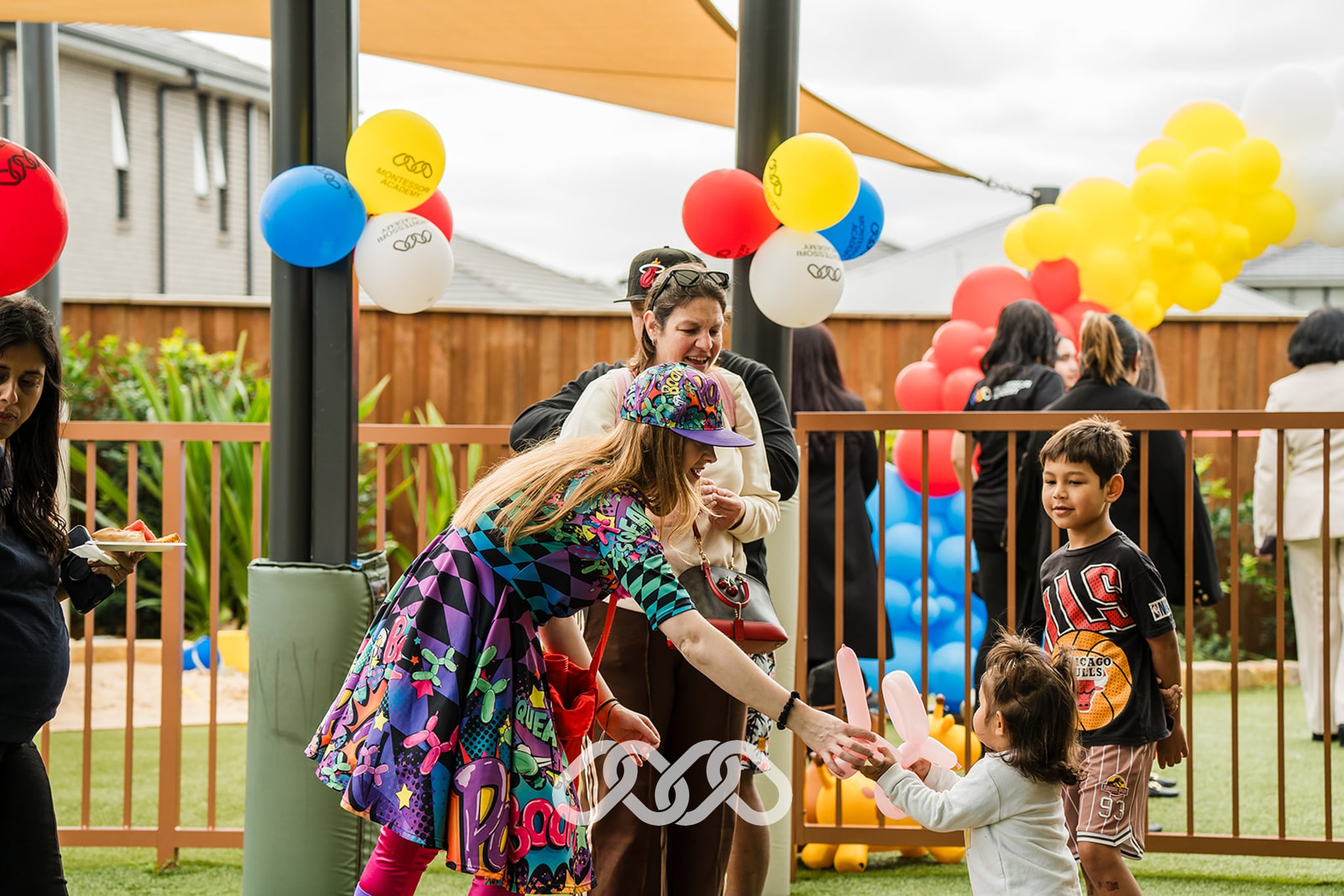balloon twister in colourful clothing passes orange balloon dog to child