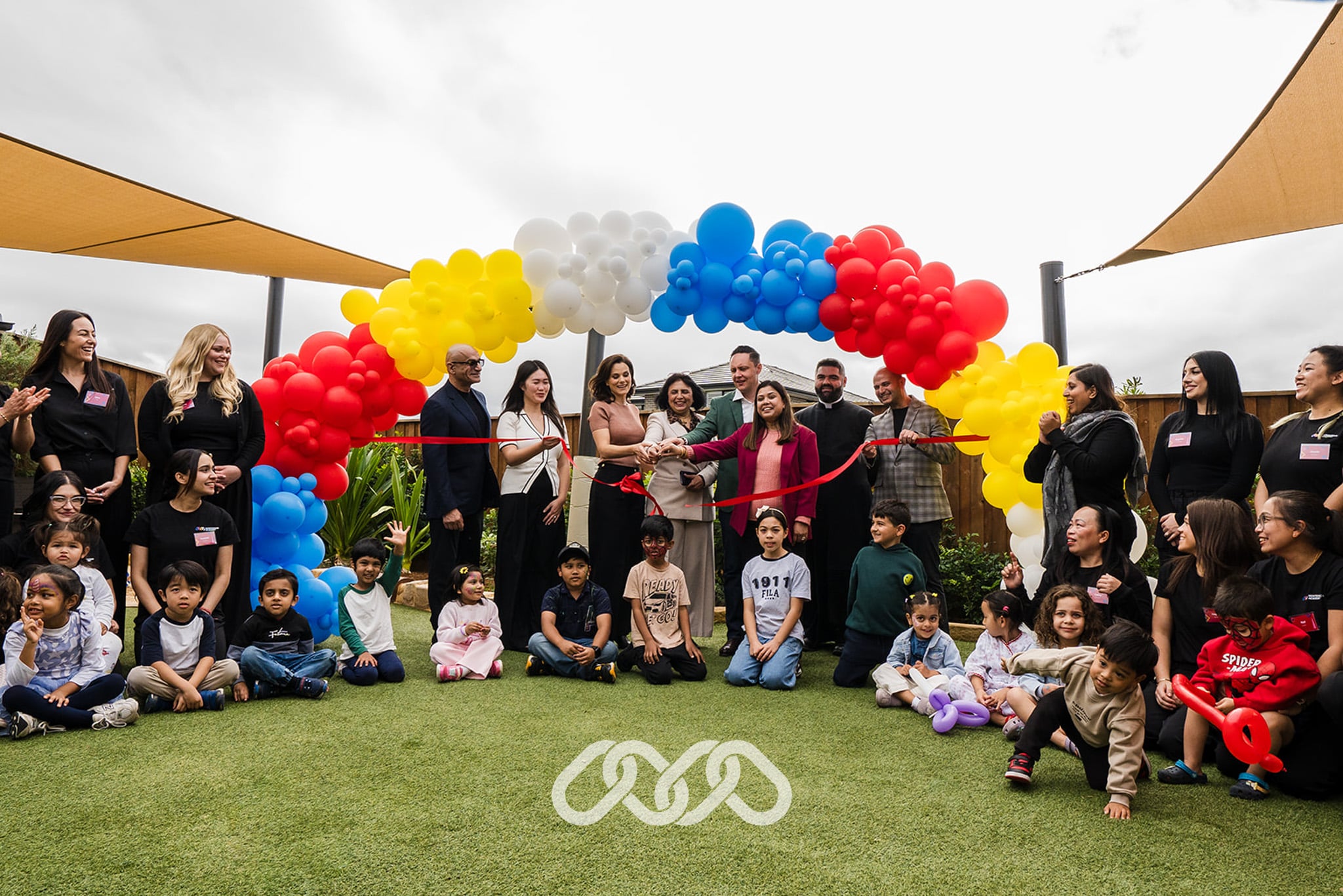 Leadership and local community leaders gather under colourful balloon arch to cut red grand opening balloon, children and montessori staff sit on either side of balloon arch celebrating the occasion