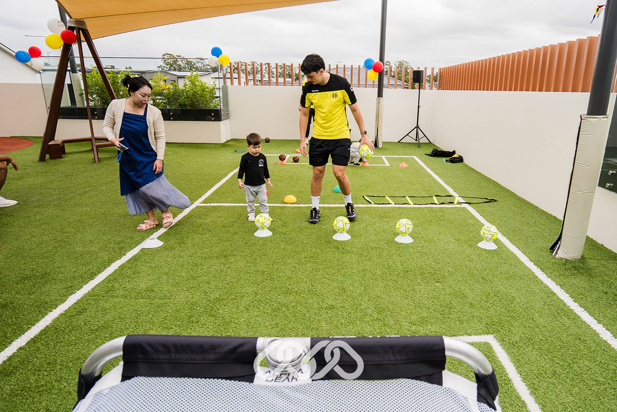 child wearing black top kicks child sized soccer ball into child sized soccer goal with assistance of sporting coach in yellow top