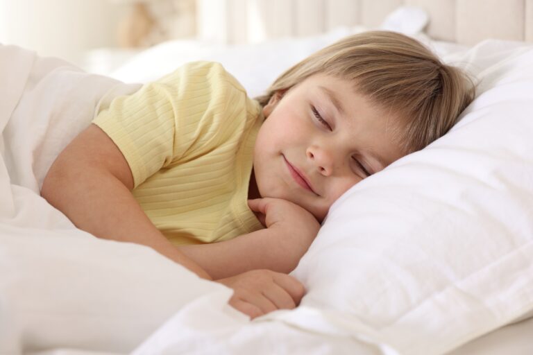 A young child in a yellow t-shirt sleeps peacefully on a white pillow, smiling, in a bright bedroom.
